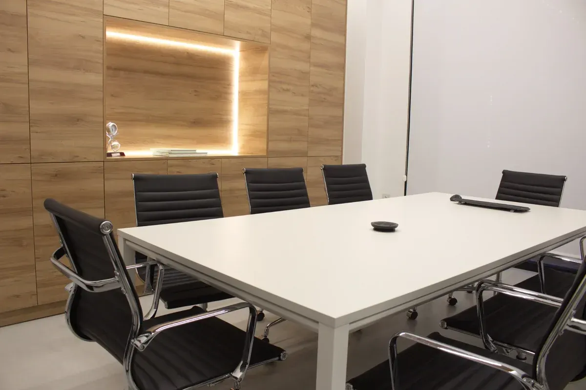 Sala de conferencias moderna con mesa blanca, sillas negras, pared revestida de madera y espejo iluminado.