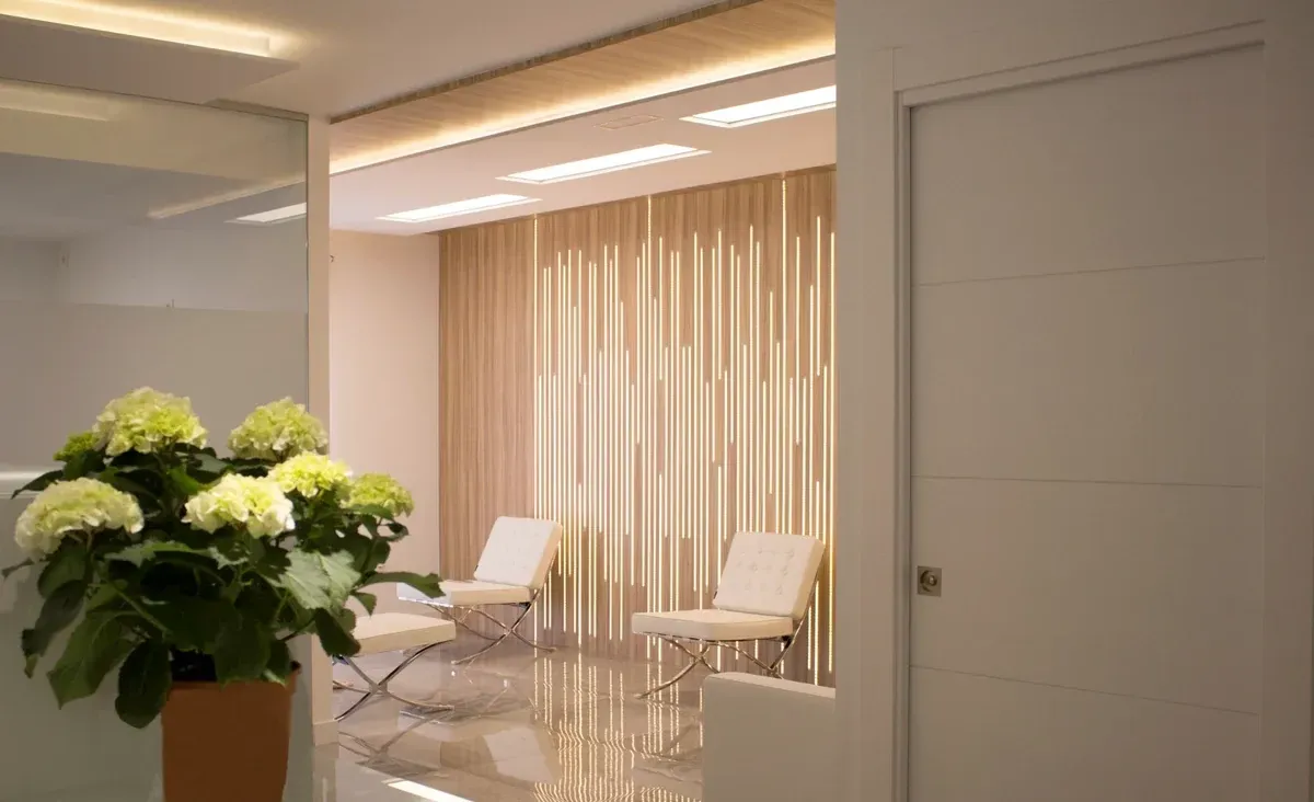 Moderna sala de espera con sillas blancas, persianas verticales y un ramo de flores blancas y verdes.
