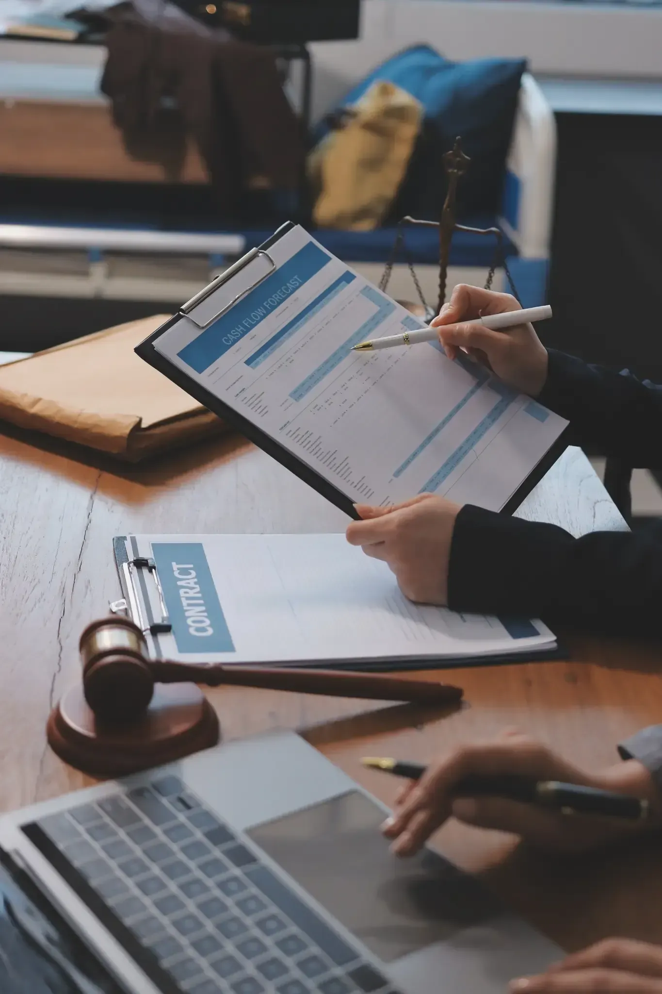 Persona señalando un documento en un portapapeles. Ordenador portátil, mazo y papeles sobre una mesa de madera.