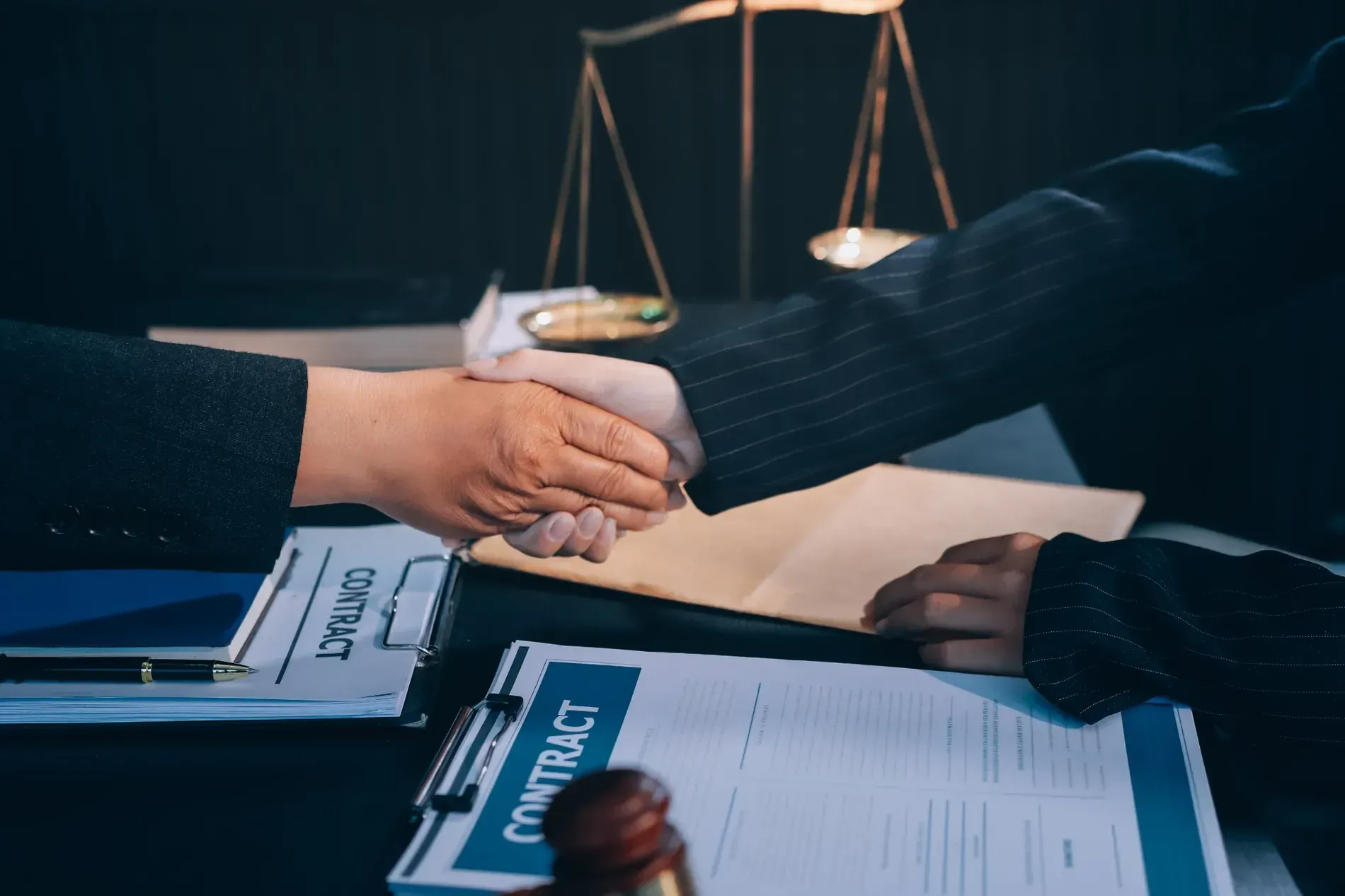 Dos personas se dan la mano sobre un contrato que está sobre una mesa; la balanza de la justicia al fondo.