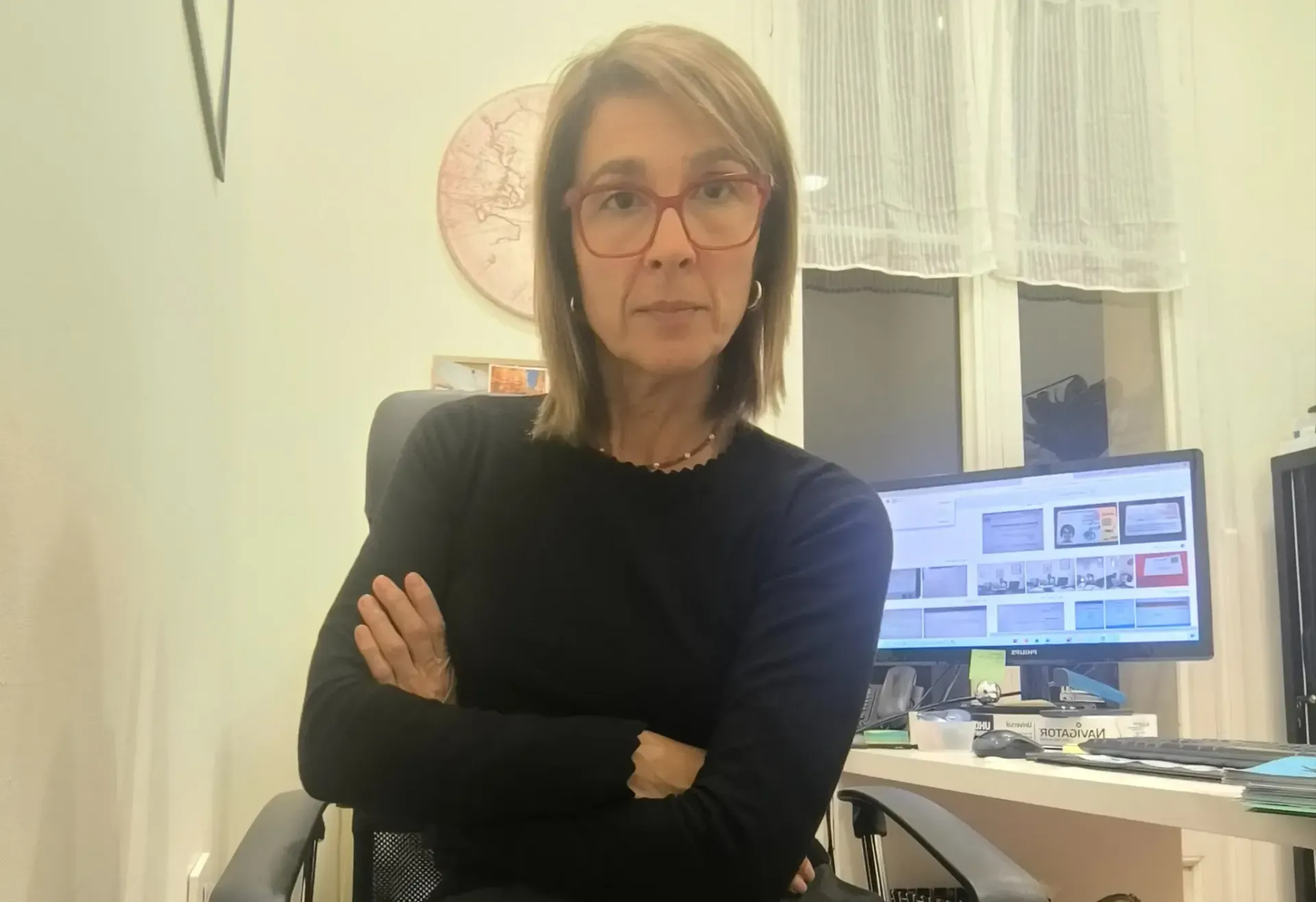Mujer con camisa negra y gafas, sentada con los brazos cruzados y la mirada al frente. Escritorio y ordenador al fondo.