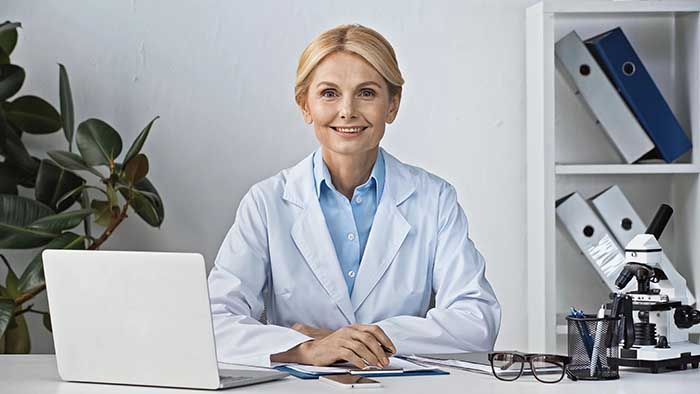 Ein Arzt im weißen Kittel sitzt in einer Büroumgebung an einem Schreibtisch mit Laptop, Dokumenten und Brille.