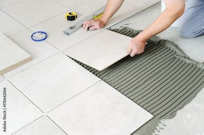 Ouvrier posant du carrelage au sol, étalant de la colle grise sous des carreaux de céramique blancs