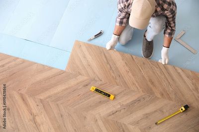 Personne posant un parquet à chevrons avec des mètres et des outils sur un sous-plancher bleu clair
