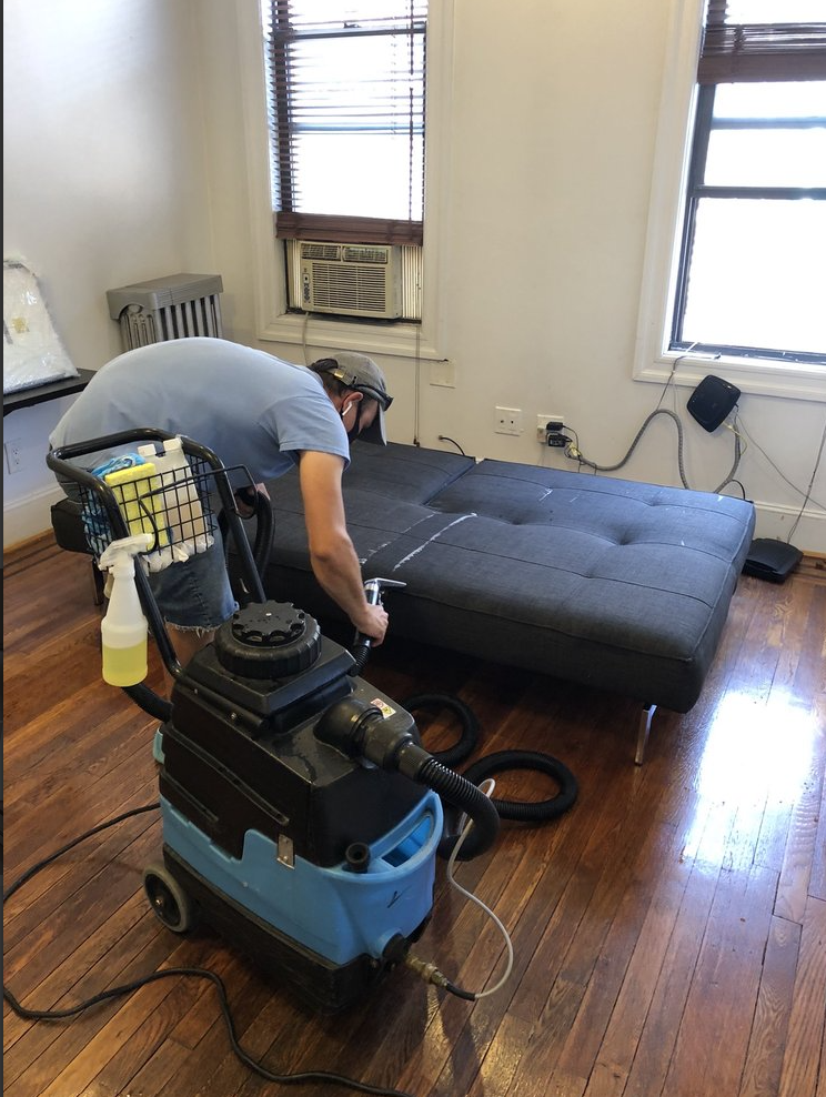Une personne passe l'aspirateur sur un canapé noir dans une pièce ensoleillée avec parquet et fenêtres.