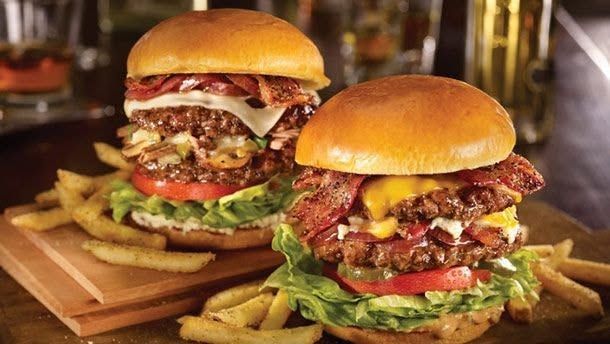 Zwei übereinandergestapelte Gourmet-Burger auf Holzbrettern mit Salat, Käse, Speck und Pommes.