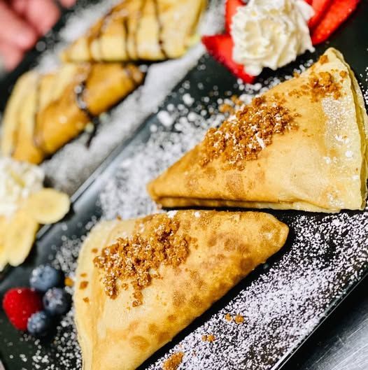 Goldbraune dreieckige Teigtaschen auf einem Tablett, mit Puderzucker bestäubt und mit Streuseln garniert, daneben Beeren und Obst