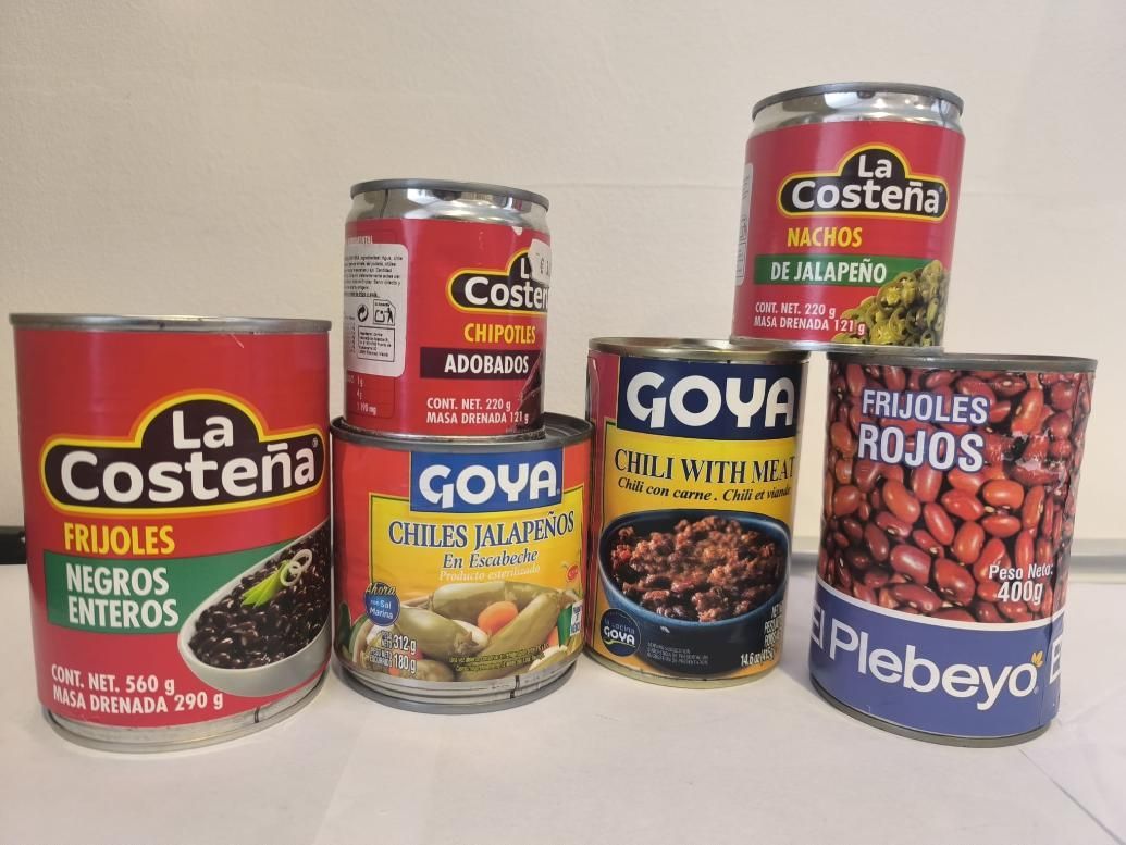 Seis latas de frijoles, en su mayoría rojos y negros, de varias marcas como Goya y La Costeña, sobre una superficie blanca.