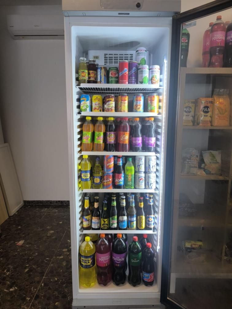 Frigorífico abastecido con diversas bebidas embotelladas y enlatadas, en un entorno de tienda.