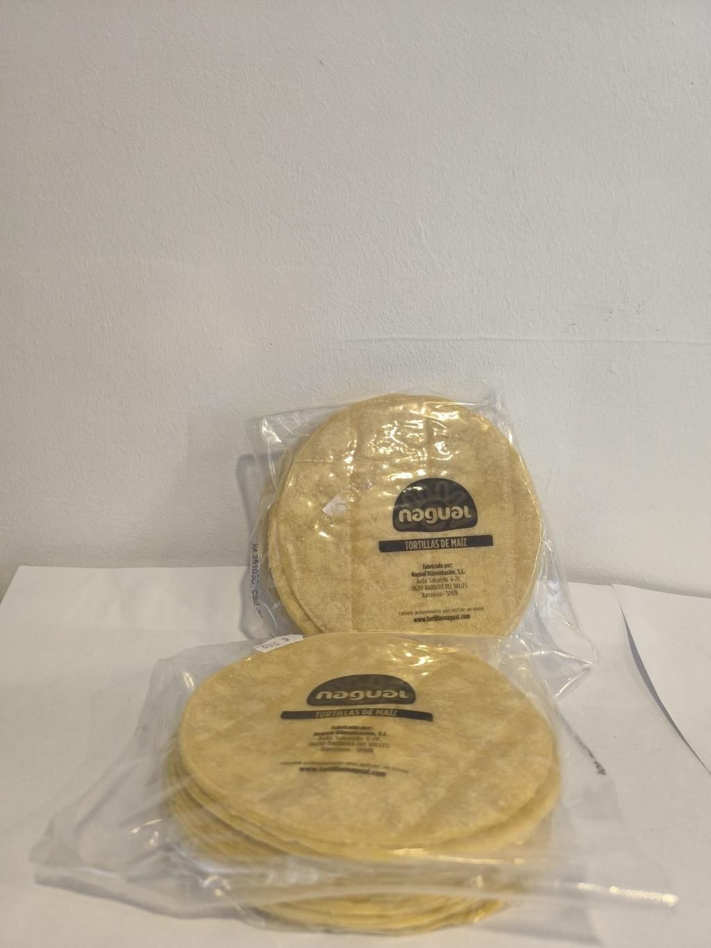 Dos paquetes de tortillas sobre una superficie blanca, contra una pared blanca.