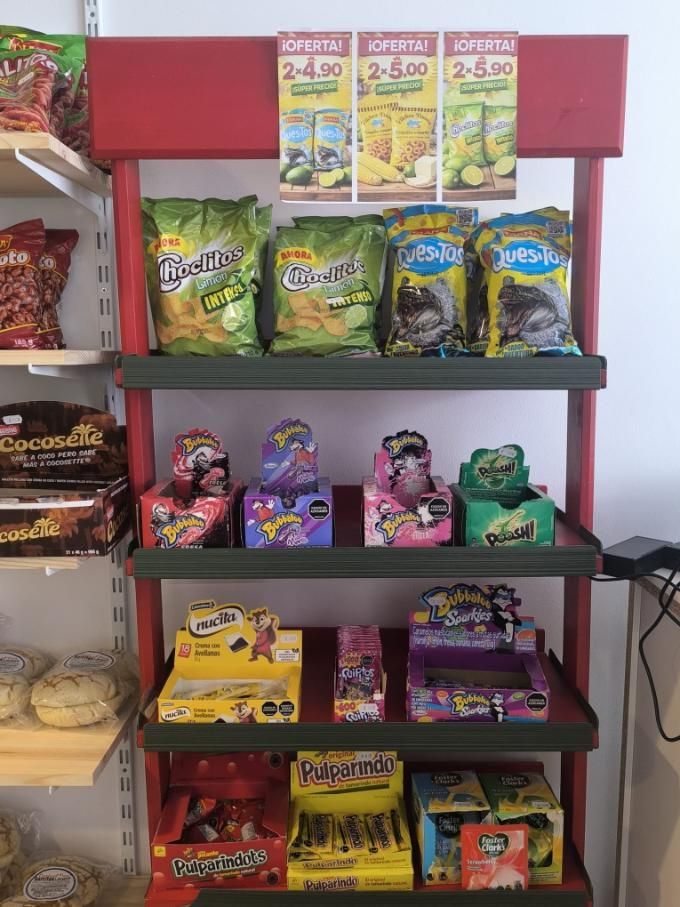 Exhibición de refrigerios con papas fritas, dulces y jugos en caja. Estantes rojos contra una pared blanca.