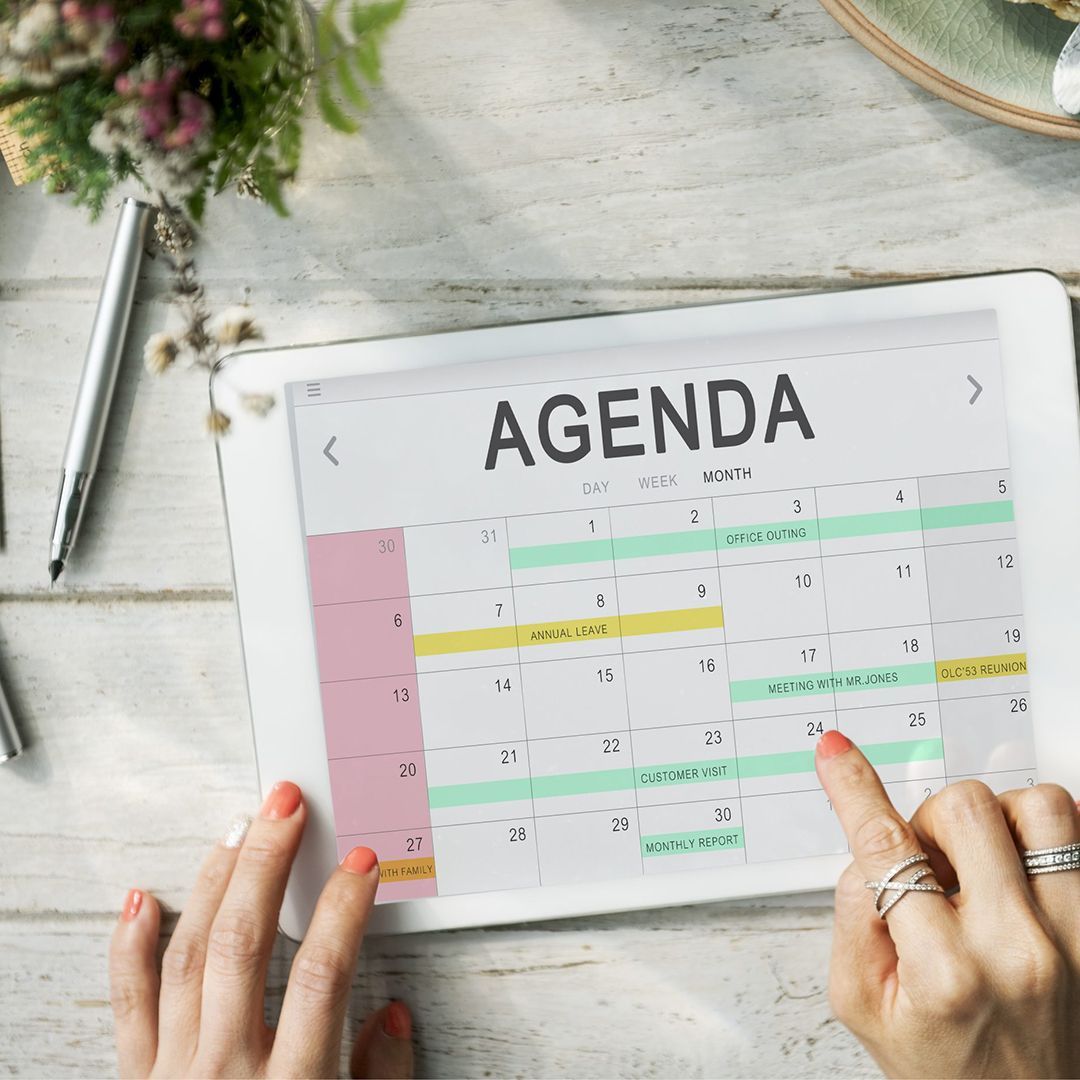 Mani che usano un tablet che mostra un calendario mensile su una scrivania bianca con fiori e una penna