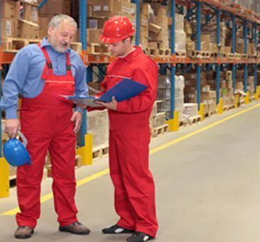 Dos hombres con overoles rojos están parados en un almacén mirando un portapapeles.