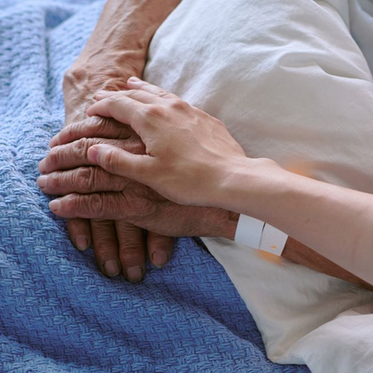 Deux mains jointes sur un lit, l'une avec un bracelet d'hôpital et une couverture blanche à proximité.