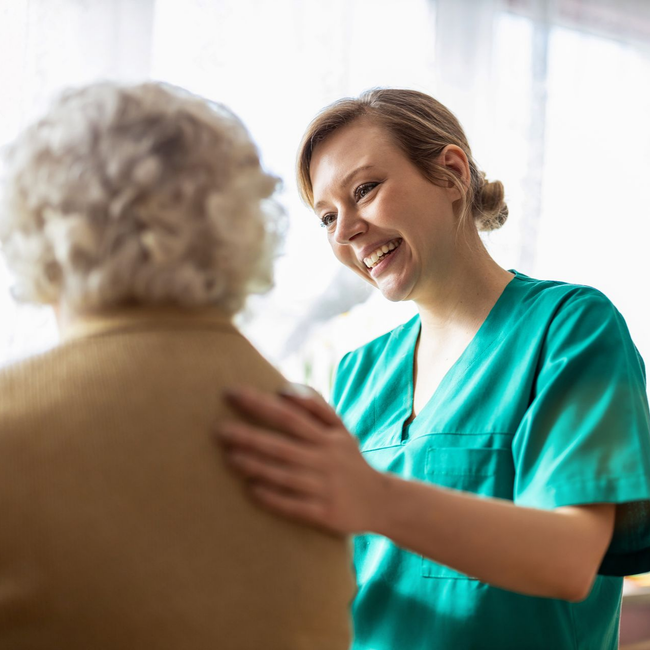 Une infirmière en blouse turquoise sourit et réconforte un patient dans un environnement de soins chaleureux et lumineux.