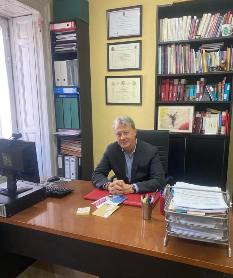El hombre en el escritorio en la oficina sonríe; estantes con libros y certificados.