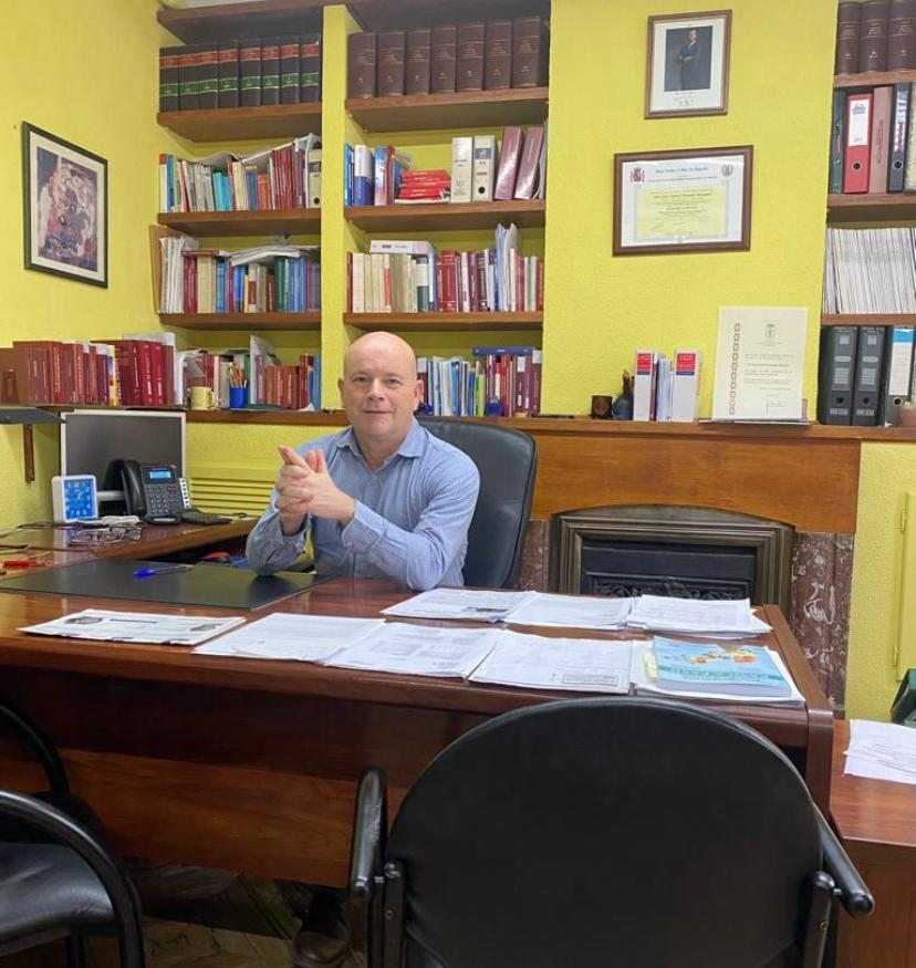 Hombre calvo con camisa azul sentado en un escritorio en una biblioteca, con documentos, estantes de libros y una chimenea.