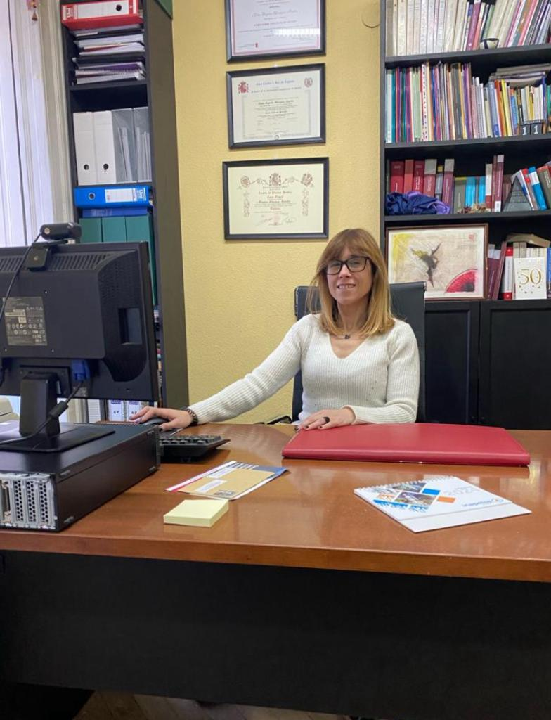 Mujer en la oficina, sonriendo, sentada en el escritorio con computadora, libros y documentos enmarcados en la pared.