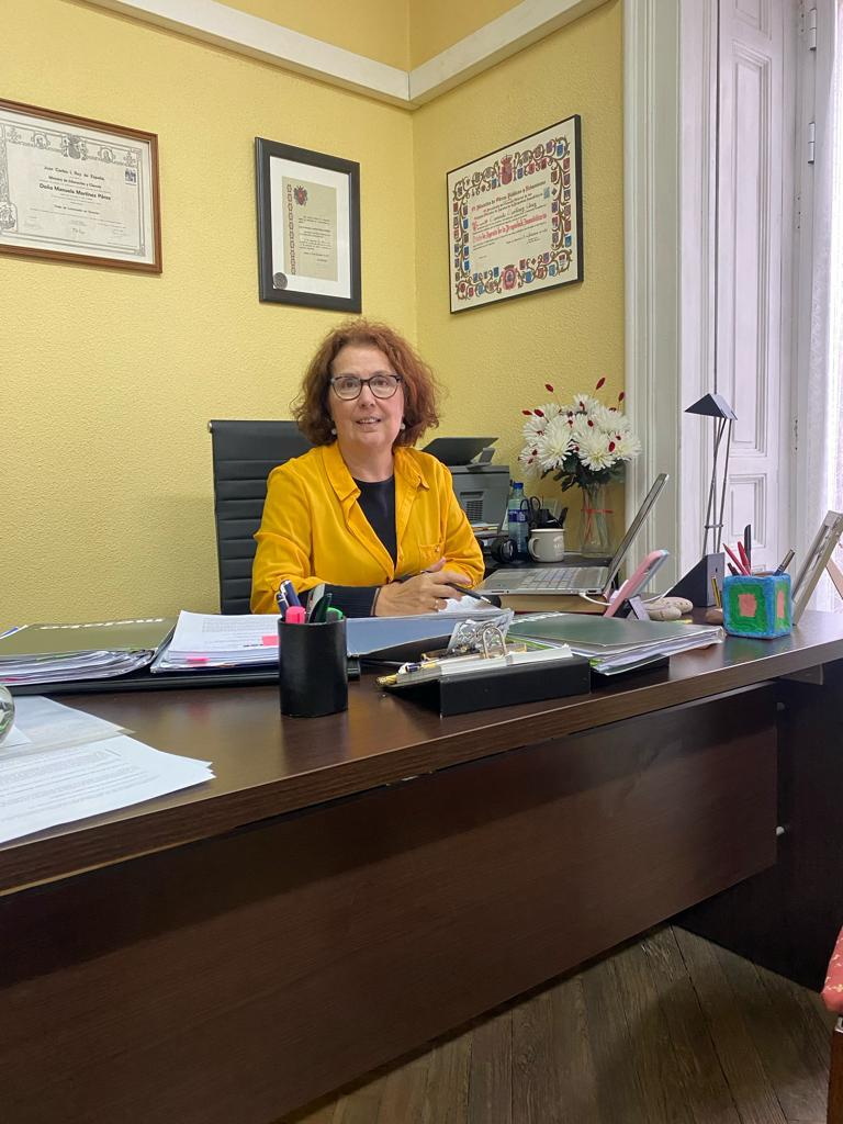 Una mujer con un blazer amarillo se sienta en un escritorio en la oficina, sonriendo.
