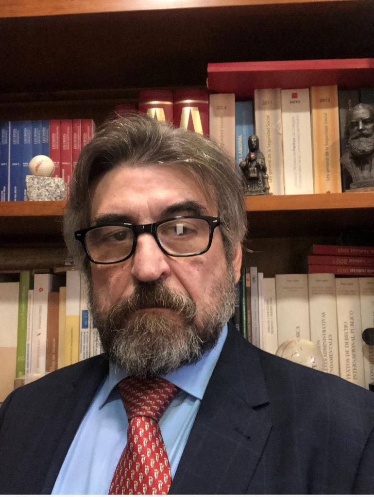 Hombre con gafas, barba y traje delante de estanterías, mirando a la cámara.