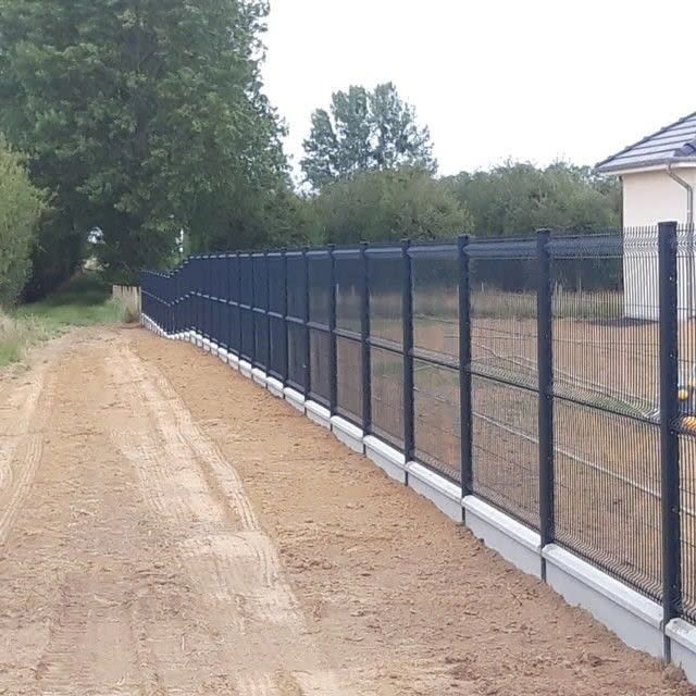 Une clôture de fer noir avec une base en béton longe un chemin de terre.