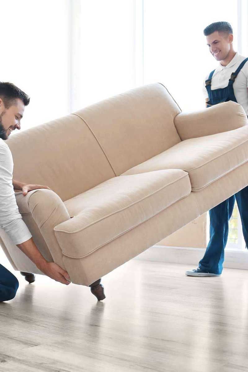 Dos operarios transportan un sofá beige a través de una habitación luminosa.