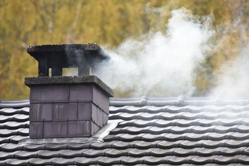 Des volutes de fumée blanche s'échappent d'une cheminée aux tuiles sombres, sur un toit de bardeaux gris, se détachant sur un fond flou aux tons dorés.