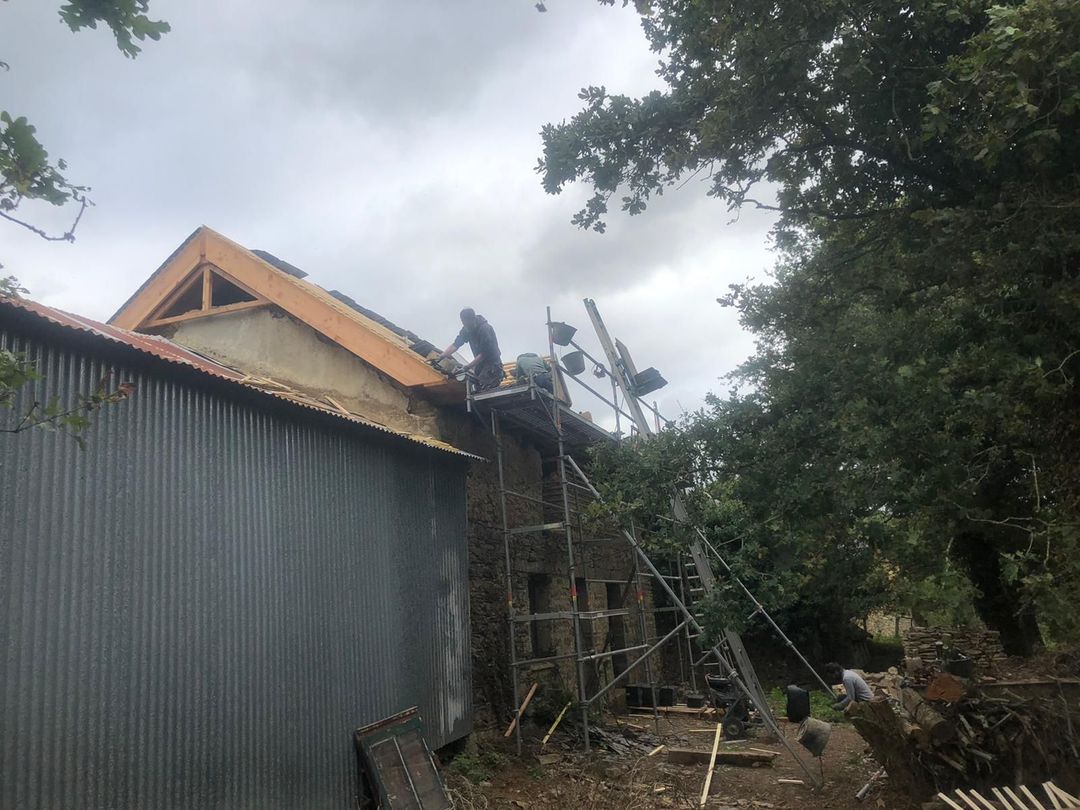 Un ouvrier, juché sur un échafaudage, répare le toit d'un bâtiment en pierre, à côté d'une structure métallique ondulée, sous un ciel nuageux.