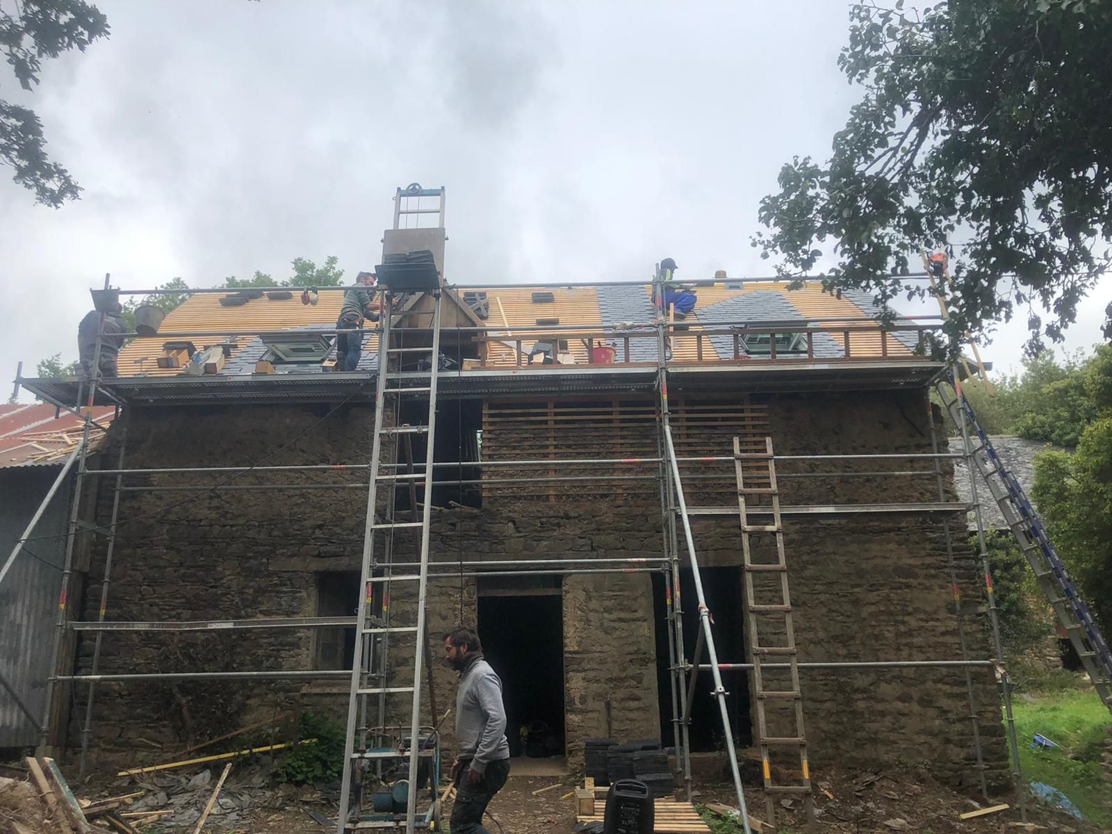 Des ouvriers effectuent des réparations sur le toit d'un vieux bâtiment en pierre, à l'aide d'échafaudages et d'échelles, sous un ciel nuageux.