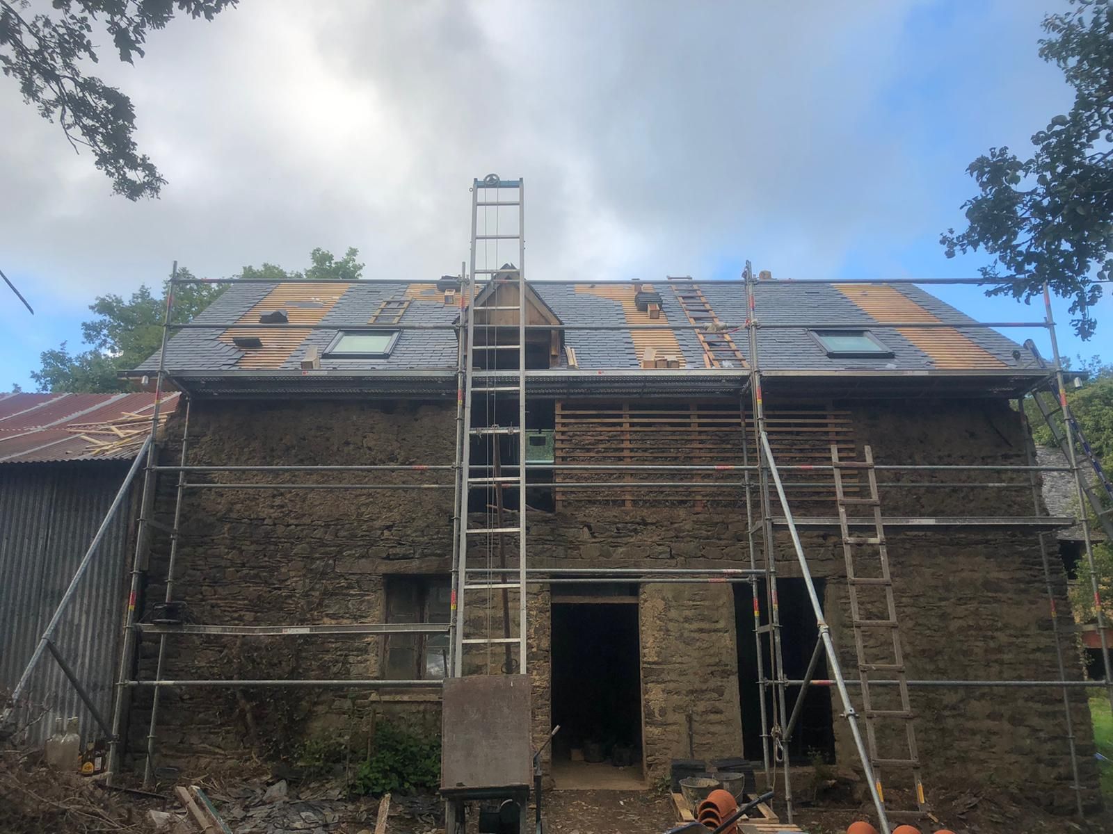 Une maison en pierre en cours de rénovation de toiture, avec des échafaudages, une échelle escamotable et de nouvelles tuiles sous un ciel bleu.