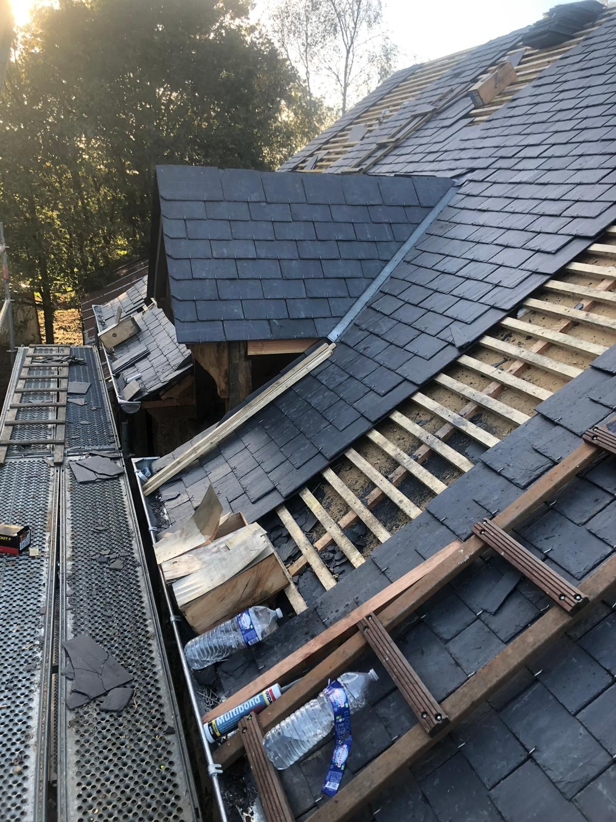 Vue en plongée d'un toit résidentiel en construction, montrant des tuiles d'ardoise foncées, des liteaux en bois et une échelle métallique.