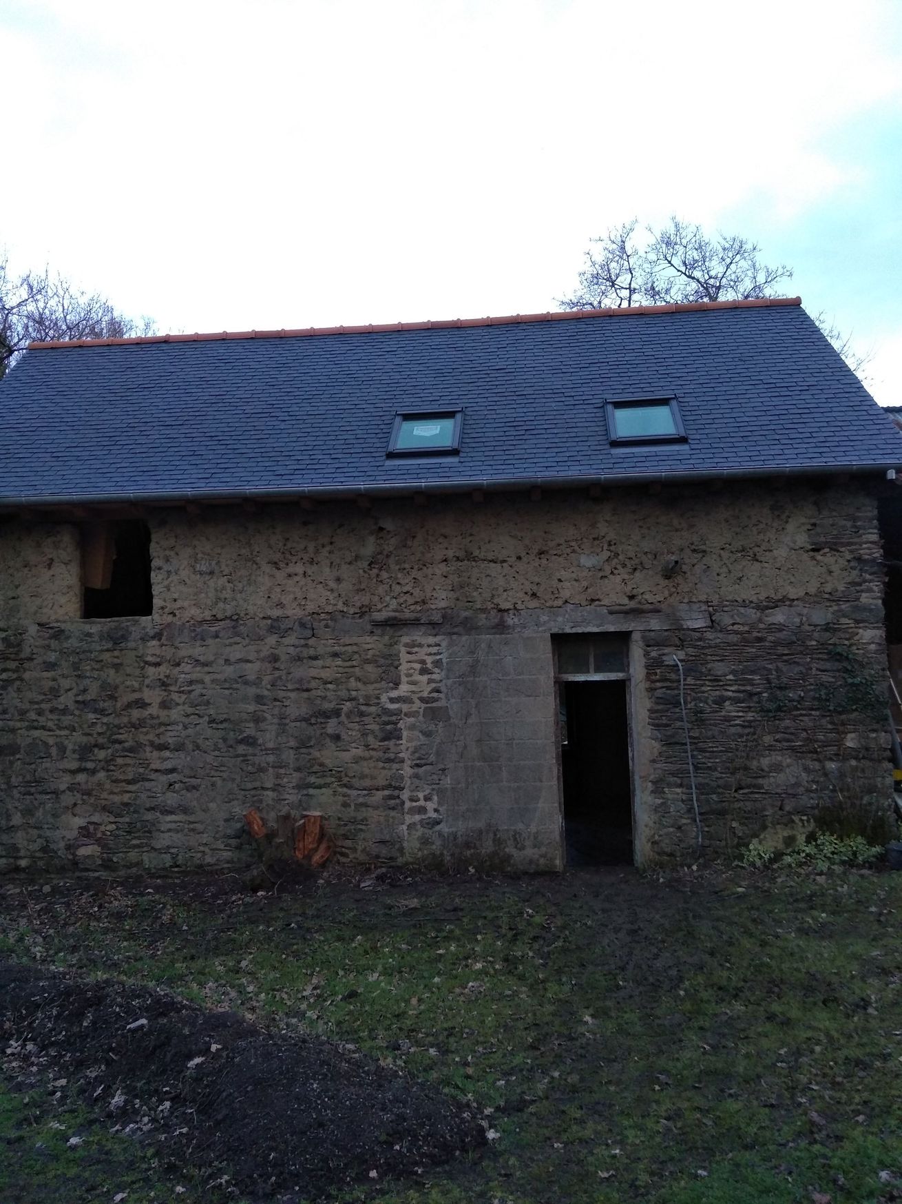 Une grange rustique en pierre à la texture patinée et au toit de bardeaux sombres à motifs incurvés, percé de deux petits puits de lumière.