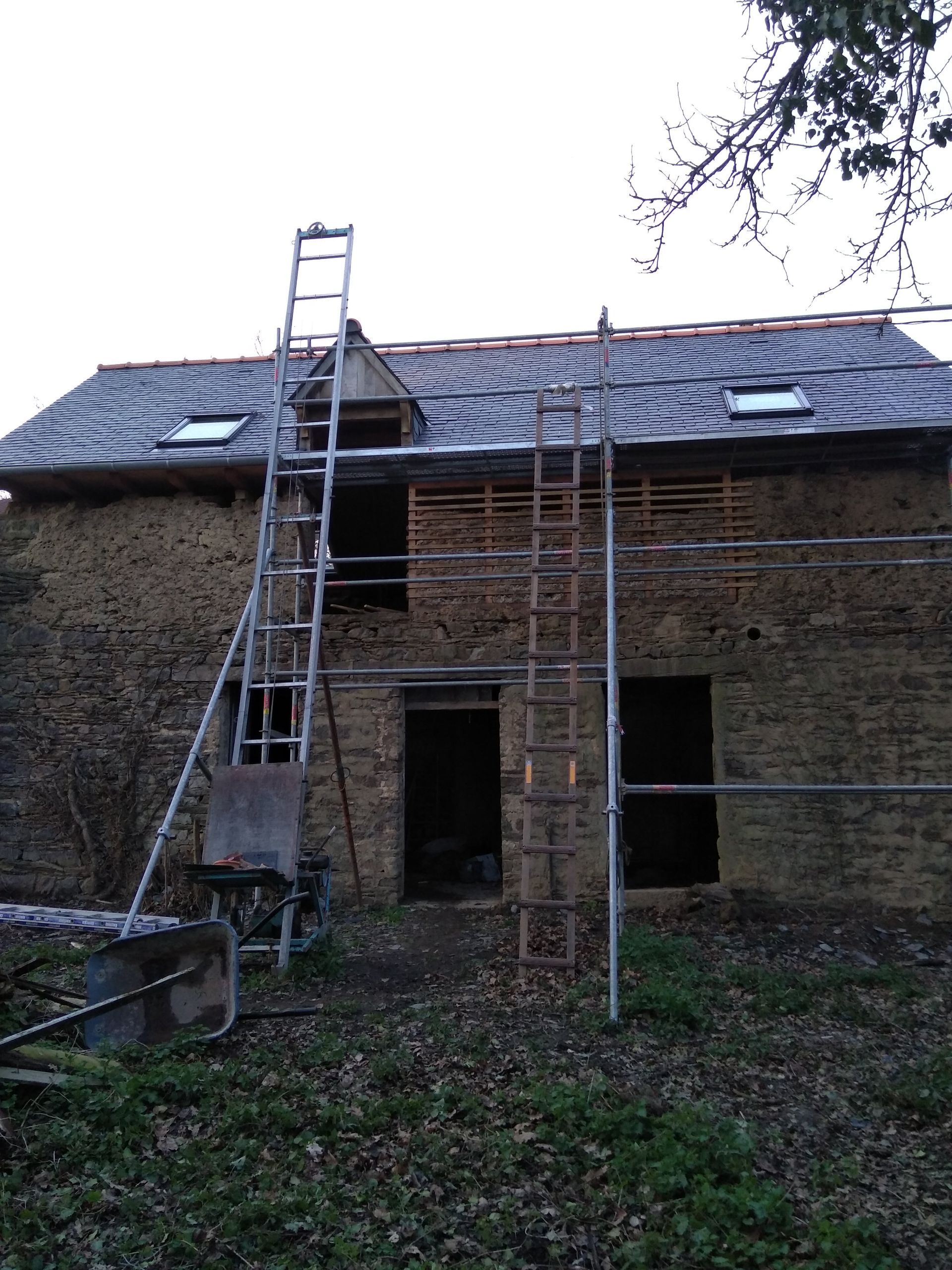 Une maison en pierre en rénovation, avec des échafaudages et des échelles adossés à l'extérieur, présentant un toit de tuiles et une porte ouverte.