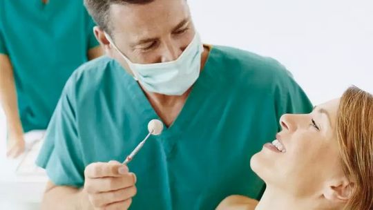 Dentista con bata verde y mascarilla examinando a un paciente sonriente con un espejo dental.