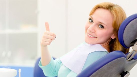 Mujer sonriente en un sillón dental azul levantando el pulgar en una luminosa sala de consulta.