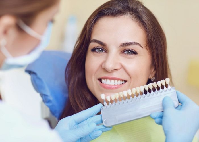 Paciente sonriente en el sillón del dentista mientras el profesional, con guantes, sostiene una guía de colores dentales cerca.