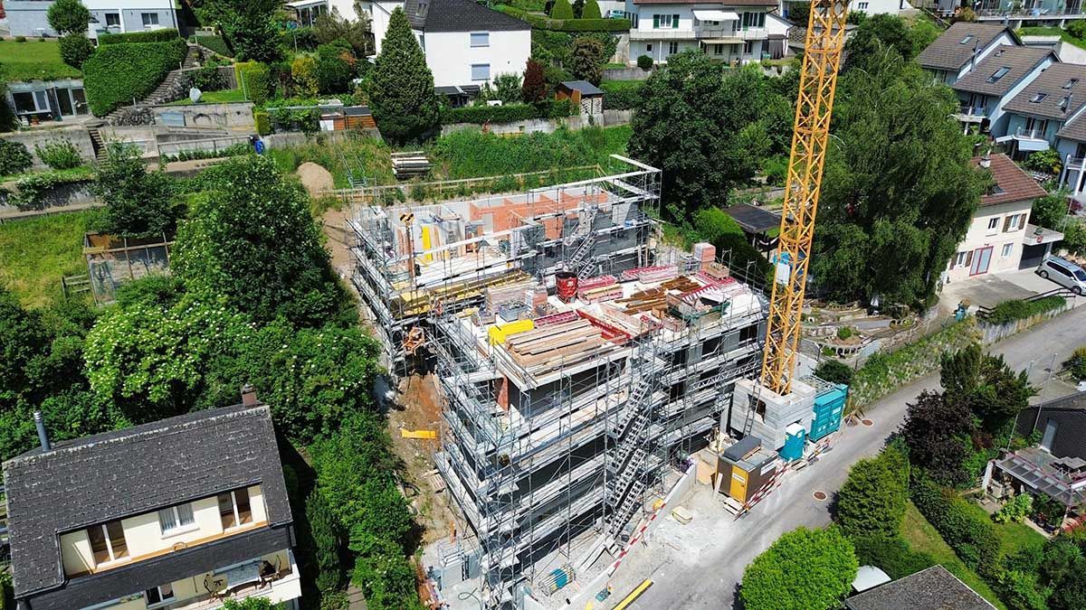 Luftaufnahme eines im Bau befindlichen mehrstöckigen Gebäudes, umgeben von Gerüsten, einem hohen gelben Kran und Bäumen.