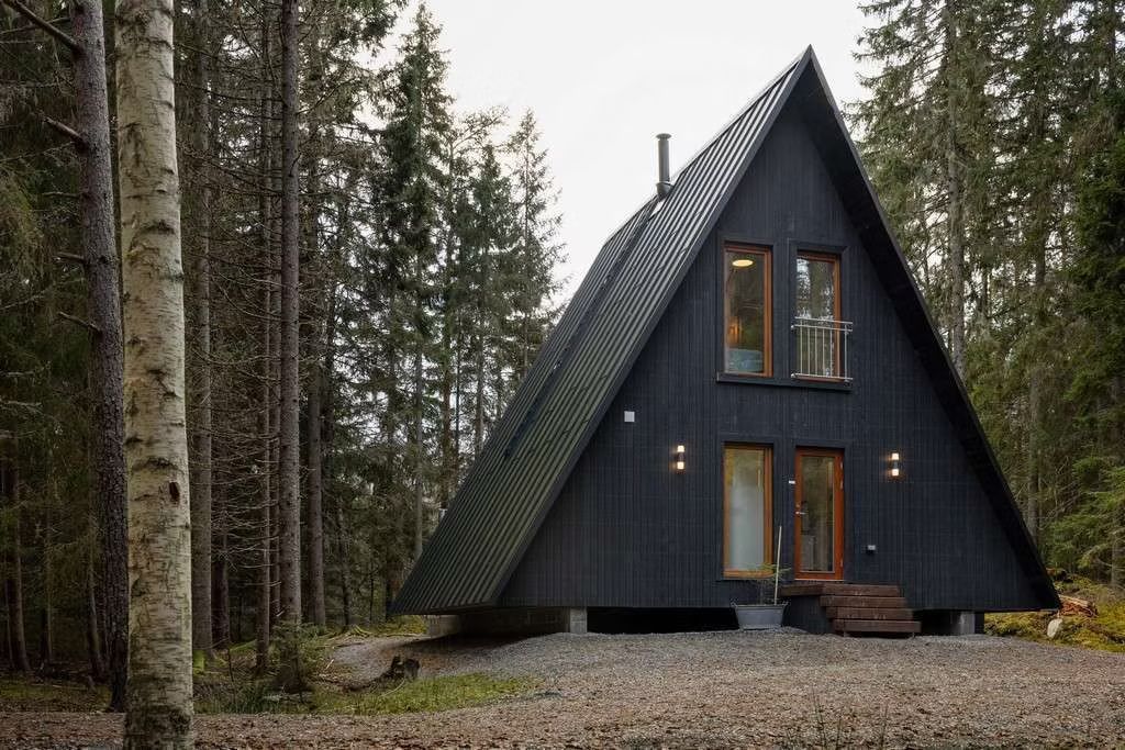 In einem dichten Wald steht eine moderne, dunkelwandige A-förmige Hütte mit hohen, schmalen Fenstern und einem Kieshof.