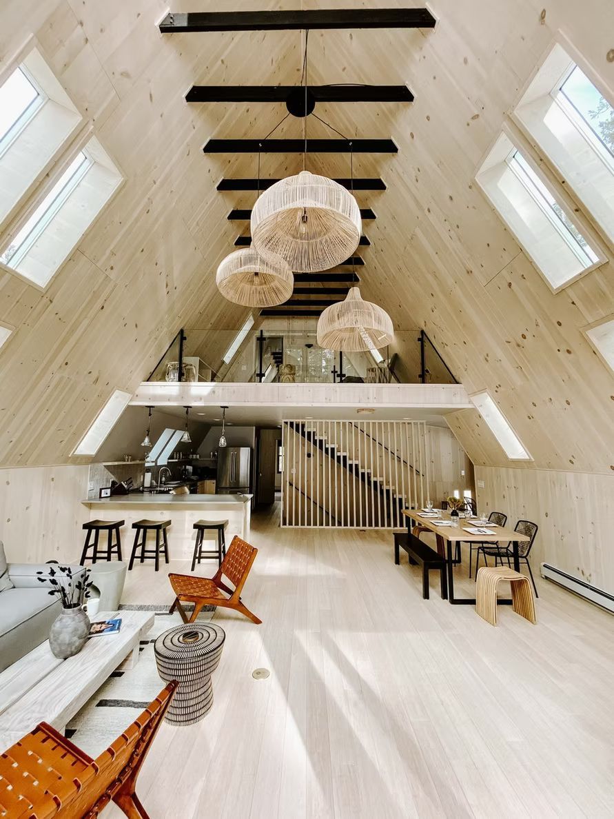 Ein modernes A-förmiges Hütteninterieur mit hellen Holzvertäfelungen, einem Schlafboden, Oberlichtern, Hängeleuchten und einem offenen Grundriss.