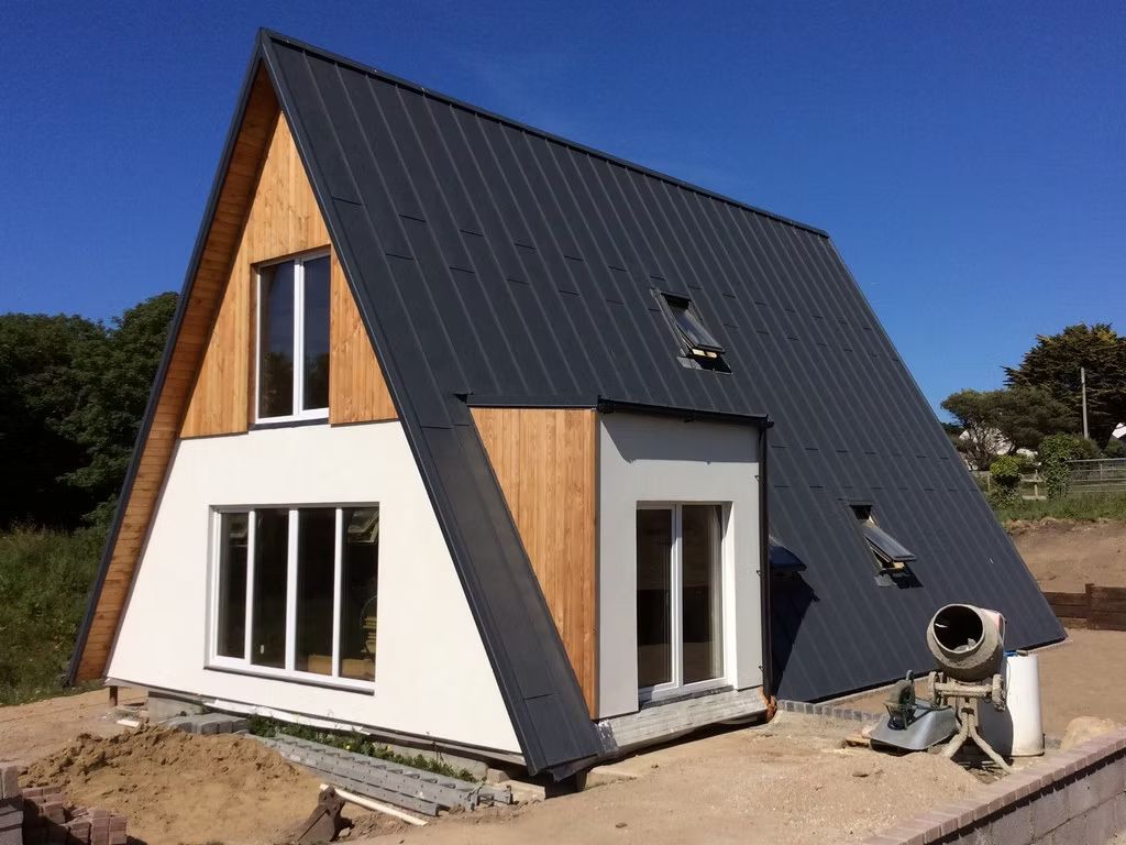 Modernes A-förmiges Haus im Bau mit dunklem Metalldach, weißen Wänden und Akzenten aus Holzverkleidung vor blauem Himmel.
