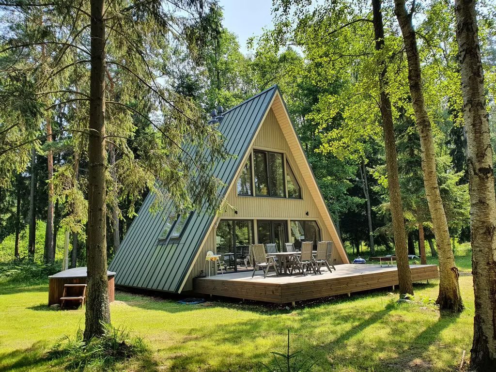 Mitten im Wald steht eine A-förmige Hütte mit grünem Dach auf einer sonnigen Holzterrasse. Sie verfügt über große Fenster und einen Whirlpool im Freien.