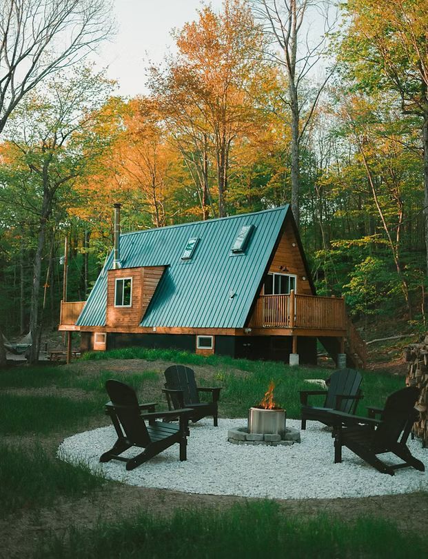 In einem Wald steht hinter einer Feuerstelle, umgeben von vier schwarzen Stühlen, eine hölzerne A-förmige Hütte mit grünem Dach.