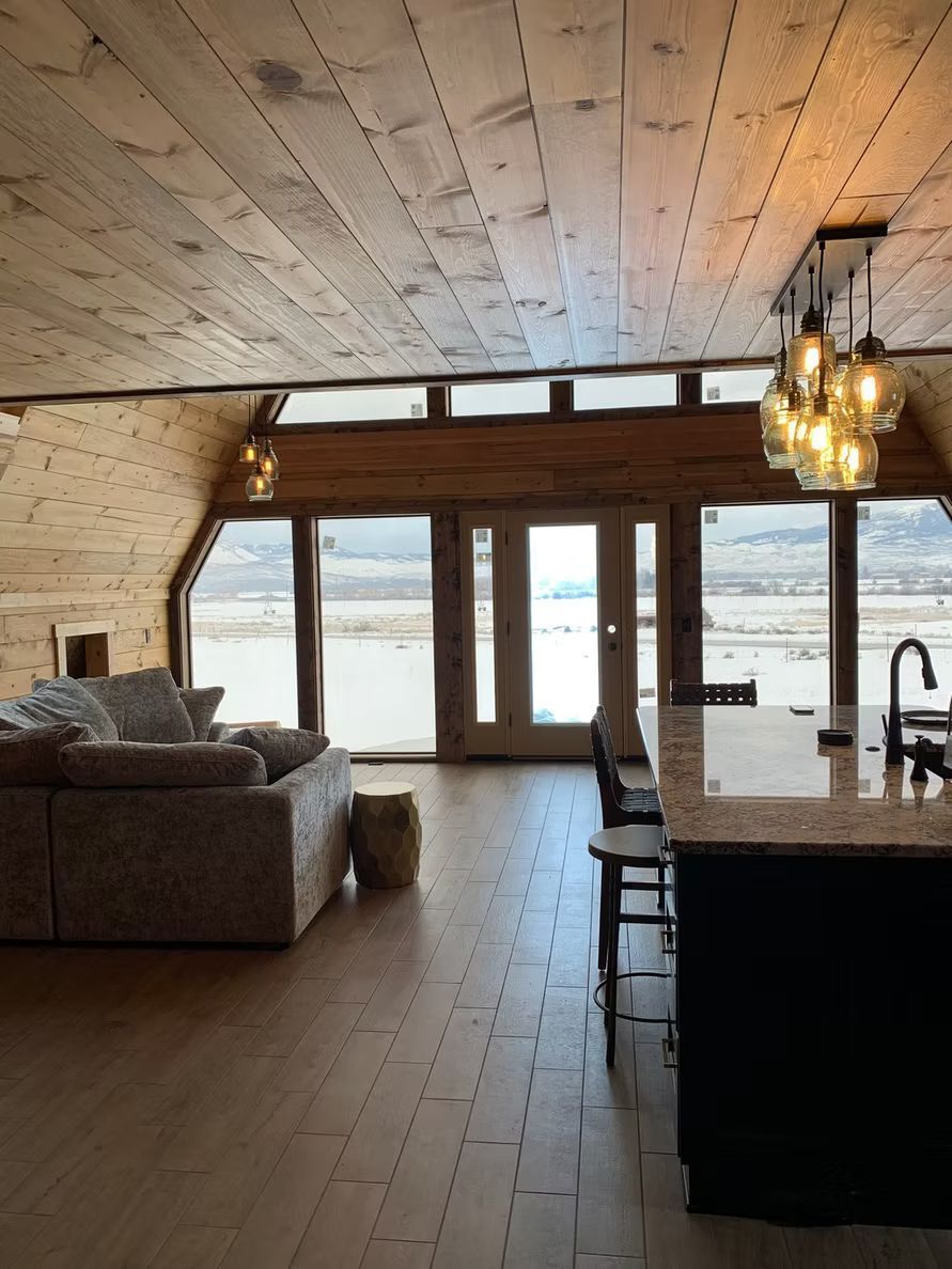 Ein modernes, offenes Hütteninterieur mit Sofa, Kücheninsel, Holzvertäfelung und großen Fenstern mit Blick auf ein Feld.