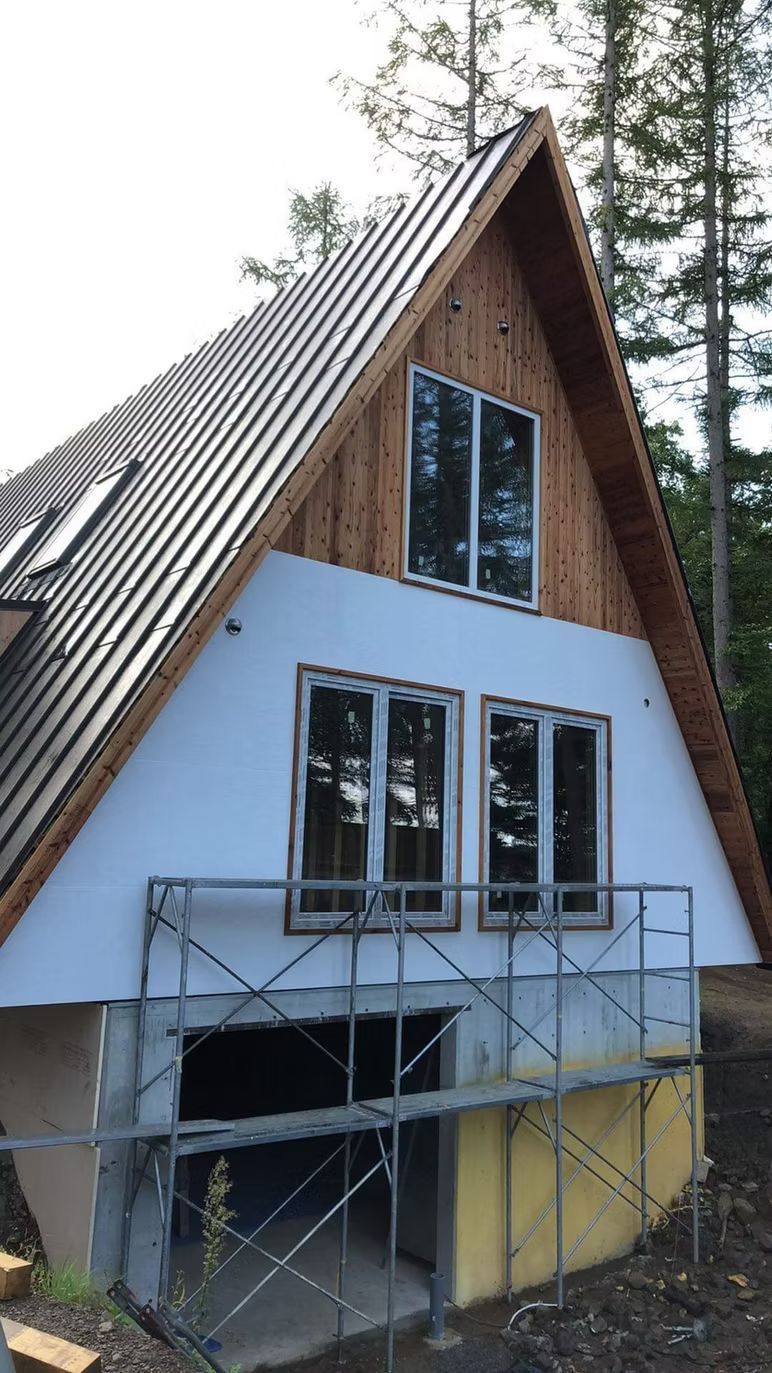 Ein dreieckiges Haus im Bau mit weißer, isolierter Fassadenverkleidung, neuen Fenstern und einem Metallgerüst davor.