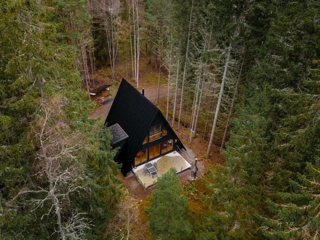 Inmitten eines üppigen, immergrünen Waldes liegt eine A-förmige Hütte mit schwarzem Dach und einer kleinen Holzterrasse.