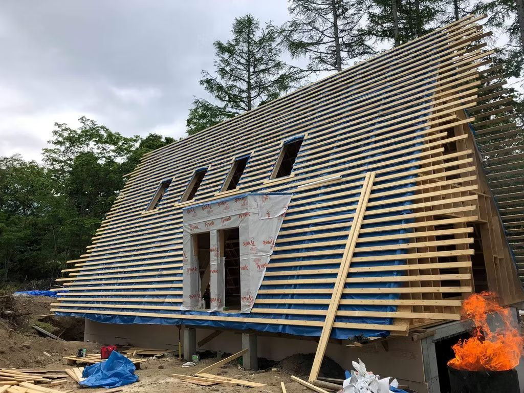 Ein modernes A-förmiges Haus im Bau, mit Holzrahmenkonstruktion, Dachlatten und Dachgaube, eingebettet in ein Waldgebiet.