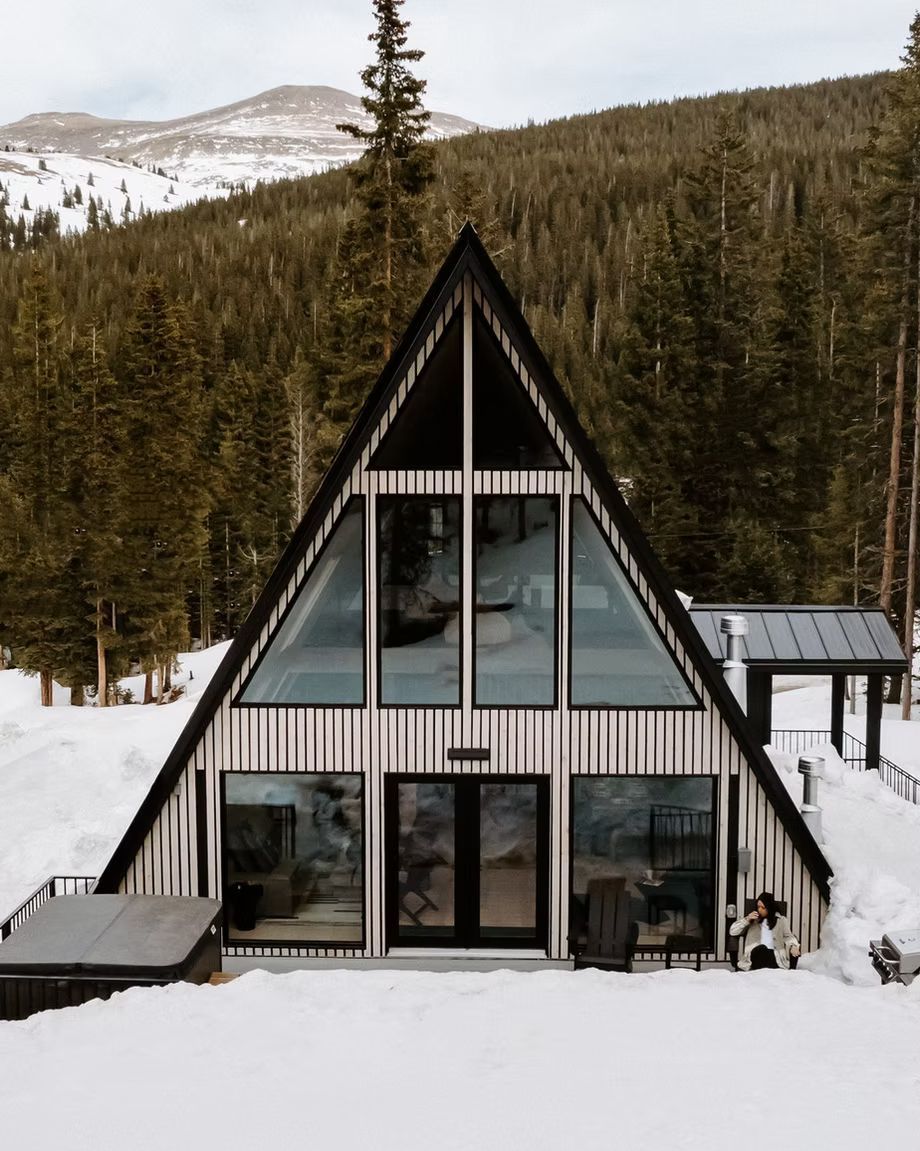 Inmitten einer verschneiten Berglandschaft, umgeben von Kiefern, liegt eine gestreifte A-förmige Hütte mit einem Whirlpool auf der Terrasse.