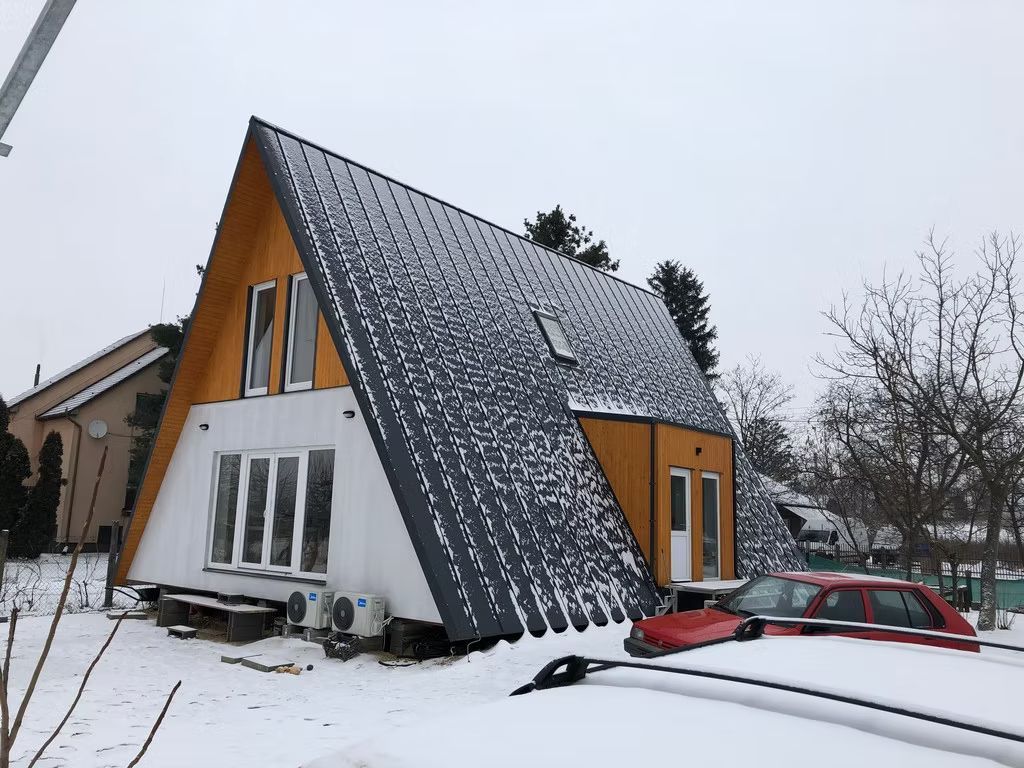 Ein modernes, weißes A-förmiges Haus mit dunklem, schneebedecktem Dach steht an einem bewölkten Tag in einem verschneiten Hof neben einem roten Auto.
