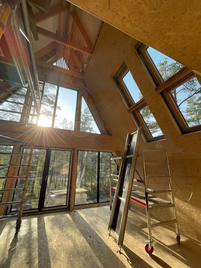 Innenansicht einer im Bau befindlichen A-förmigen Konstruktion mit OSB-Wänden, großen Fenstern und einem beweglichen Gerüst in der Mitte.