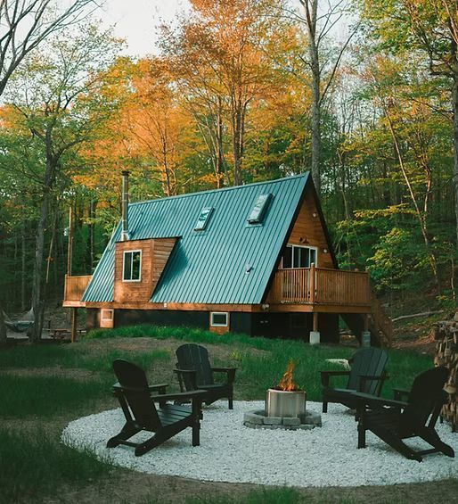 In einem Wald steht eine Holzhütte mit blauem Metalldach, auf der sich eine Feuerstelle mit vier Stühlen auf einer Kiesterrasse befindet.